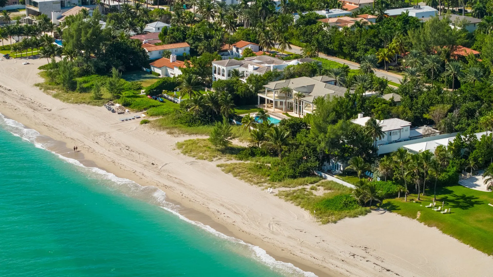 Aerial view of a beachfront with several houses nestled among trees. The sandy beach meets turquoise water, with a few people walking and beach chairs set up near the shore.